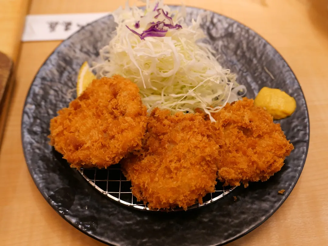A stunning close-up of a golden-brown crispy pork cutlet, perfectly fried and glistening with oil, resting on a bed of vibrant green cabbage, with a dipping sauce alongside
